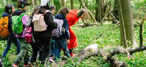 A group of children are in the woods with an actress dressed as Little Red Riding Hood. They are wearing headphones and looking at a fallen tree trunk.