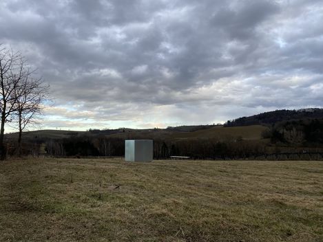 Landschaft mit hügeligem Grasland, einem kahlen Baum links, einem kleinen grauen Kubus in der Mitte und bewölktem Himmel.