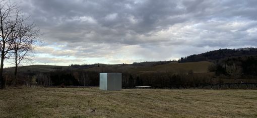 Landschaft mit hügeligem Grasland, einem kahlen Baum links, einem kleinen grauen Kubus in der Mitte und bewölktem Himmel.