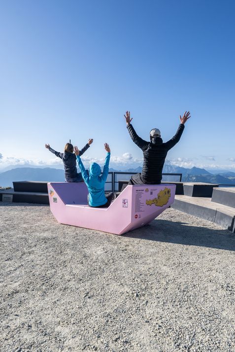 Three people sit on a pink Enzi enjoying the panoramic view from Mount Schafberg.