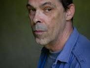 Portrait of a person with dark hair wearing a blue shirt against a green background.