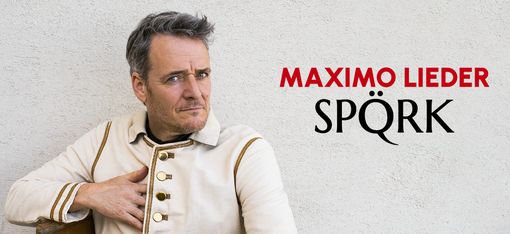 Ein Mann in einer historischen weißen Uniform sitzt auf einem Holzstuhl vor einer grauen Wand.