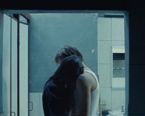 Two people embrace in a dark room in front of a window showing a gray building facade