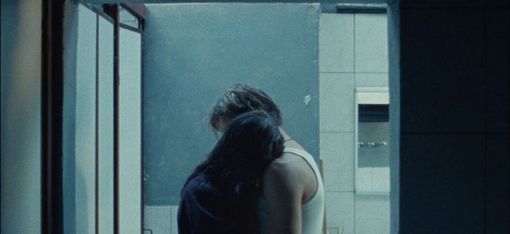 Zwei Personen umarmen sich in einem dunklen Raum vor einem Fenster mit Blick auf eine graue Gebäudefassade