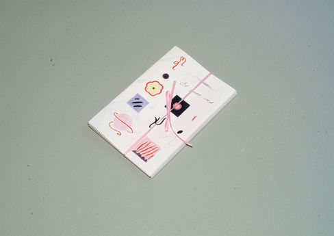 Kleines gebundenes Heft mit Zeichnungen und Mustern auf dem Umschlag, mit einem rosa Band zusammengebunden, auf einer glatten Oberfläche liegend.