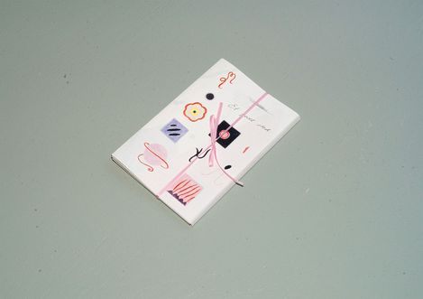 Kleines gebundenes Heft mit Zeichnungen und Mustern auf dem Umschlag, mit einem rosa Band zusammengebunden, auf einer glatten Oberfläche liegend.