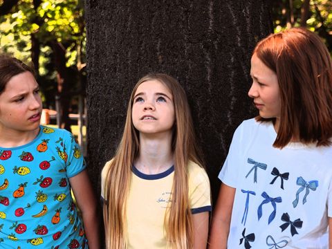Three girls are standing in front of a tree. The girl in the middle is gazing dreamily upwards, whilst the other two are looking at her questioningly.