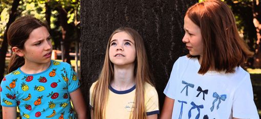 Three girls are standing in front of a tree. The girl in the middle is gazing dreamily upwards, whilst the other two are looking at her questioningly.