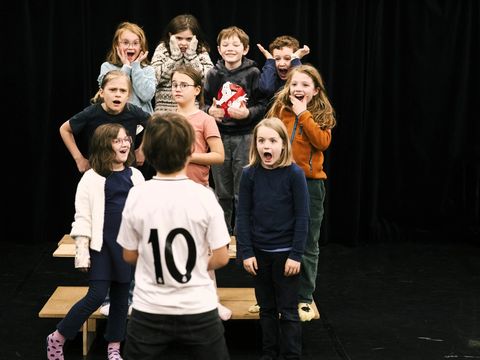 In the photo, nine children are standing on a small platform. They are reacting with different facial expressions to a child standing in front of them.