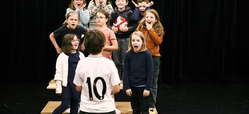 In the photo, nine children are standing on a small platform. They are reacting with different facial expressions to a child standing in front of them.