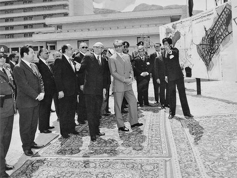 Ca. 15 Männer in dunklen Anzügen und Uniformen bekommen ein Projekt präsentiert. Sie stehen auf Perserteppichen.