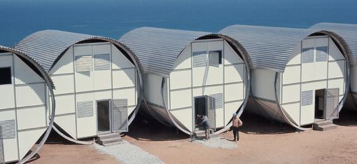 Drei liegende, tonnenähnliche Gebäude. Im Hintergrund das Meer