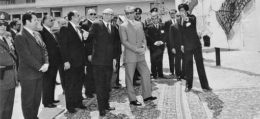 Ca. 15 Männer in dunklen Anzügen und Uniformen bekommen ein Projekt präsentiert. Sie stehen auf Perserteppichen.