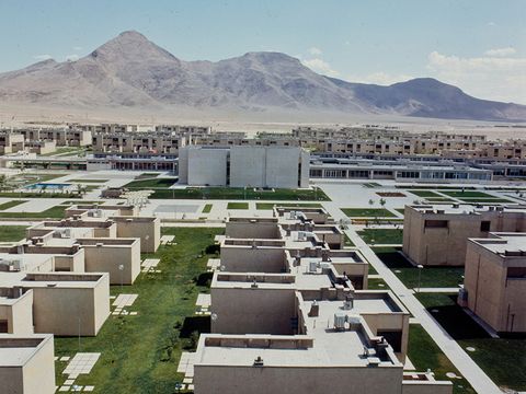 You can see a neighborhood of townhouses, with green meadows in between. A little further back, the buildings are taller. In the background, you can see a mountainous landscape.