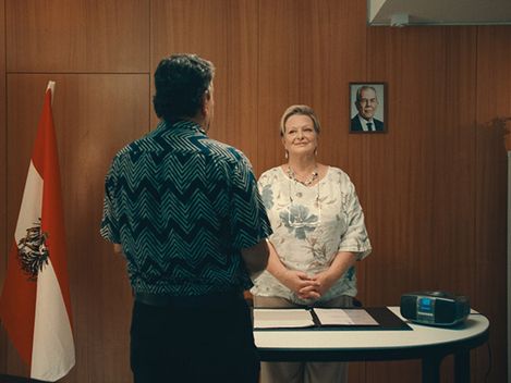 In einem, mit Holz getäfelten Raum, stehen 2 Personen. Die Frau sieht zu uns. Sie trägt ein helles Kleid. Hinter ihr hängt ein Bild von unserem Bundespräsidenten Alexander van der Bellen. Rechts neben ihr steht ein Kasten, links eine rot-weiß-rote Österreichfahne. Vor ihr auf einem weißen Tisch liegen eine aufgeschlagene Mappe und ein Radiorekorder. Ihr gebenüber, steht ein Mann, mit dem Rücken zu uns, mit gefalteten Händen.