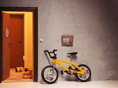 Ein gelbes Kinderrad lehnt neben einer offenen Wohnungstür. 
Aus der Wohnung dringt Licht, eine weitere Tür ist zu sehen und Spielzeug liegt am Boden.
