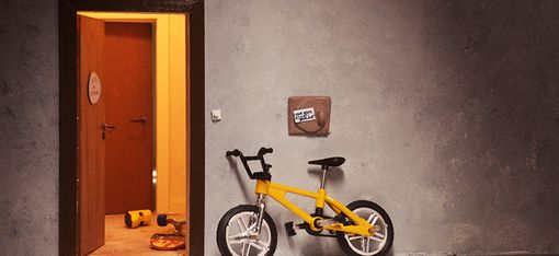 Ein gelbes Kinderrad lehnt neben einer offenen Wohnungstür. 
Aus der Wohnung dringt Licht, eine weitere Tür ist zu sehen und Spielzeug liegt am Boden.