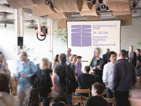 In the background is a screen displaying the text: architekturtage.at 2026
In the foreground, about 20 people are standing and sitting, engaged in lively conversation.
