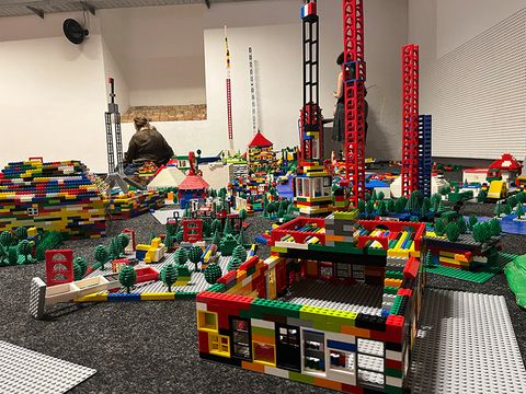 You can see a large Lego city with low and very tall buildings inside a room. In the background, there is a white wall and two people.
