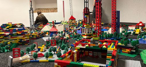 Man sieht eine gorße Legostadt mit niedrigen und sehr hohen Häusern in einem Innenraum. Im Hintergrund eine weiße Wand und zwei Personen.
