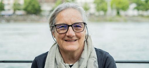 Im Vordergrund sitz eine Frau mit kurzen Haaren und einer Brille, Barbara Buser. Im Hintergrund fließt ein Gewässer und am anderen Ufer stehen einige Häuser.