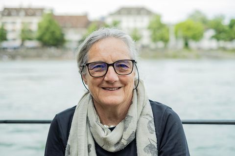 Im Vordergrund sitz eine Frau mit kurzen Haaren und einer Brille, Barbara Buser. Im Hintergrund fließt ein Gewässer und am anderen Ufer stehen einige Häuser.
