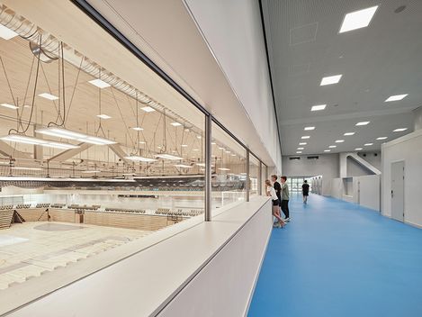 On the right side of the picture is a long, high hallway with a light blue floor. The room is divided by a wall of windows offering a view of a gymnasium on the left side of the picture.