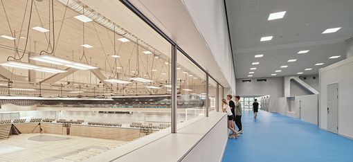 Rechts im Bild verläuft ein langer, hoher Gang mit hellblauem Fußboden. Geteilt wird der Raum durch ein Fensterwand mit Sicht auf einen Turnsaal auf der linken Bildhälfte.