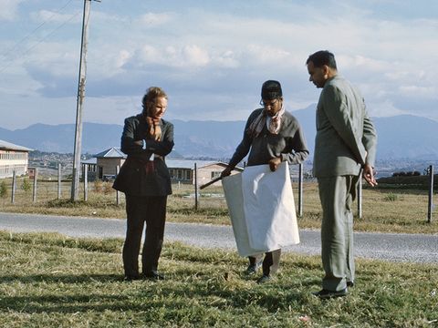 Three people are standing in a meadow. The person in the middle is holding a large piece of paper. In the background, you can see a path, a house, mountains and a cloudy sky.