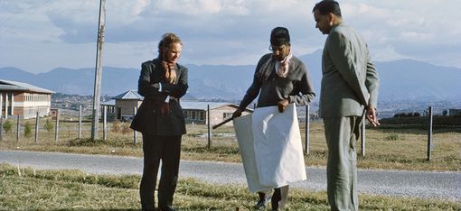 Three people are standing in a meadow. The person in the middle is holding a large piece of paper. In the background, you can see a path, a house, mountains and a cloudy sky.