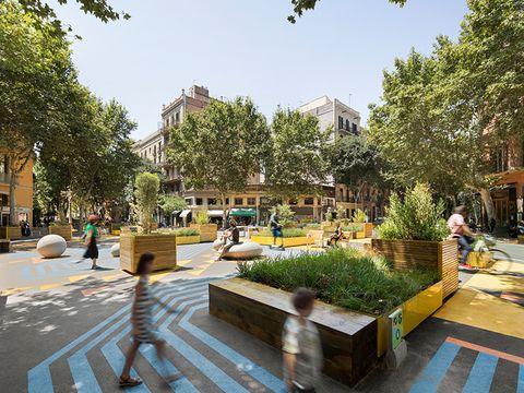 Several people are gathered in a beautiful, newly created square. Patterns on the ground break up the large open space, with several seating areas dotted amongst them. There are large planters containing trees and greenery. A residential building can be seen in the background.
