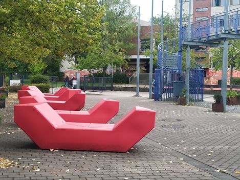 Rote Enzis stehen auf einem gepflasterten Platz vor Wohngebäuden und einer blauen Wendeltreppe, umgeben von Bäumen im urbanen Umfeld.