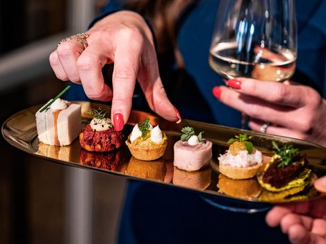 Hand mit roten Fingernägeln nimmt ein kleines Häppchen von einem goldfarbenen Tablett mit verschiedenen mundgerechten Häppchen; in der anderen Hand hält die Person ein Glas Weißwein.
