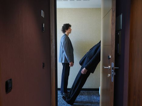 Blick durch die offene Tür eines Hotelzimmers. Ein Performer steht aufrecht, der andere scheint nach hinten z