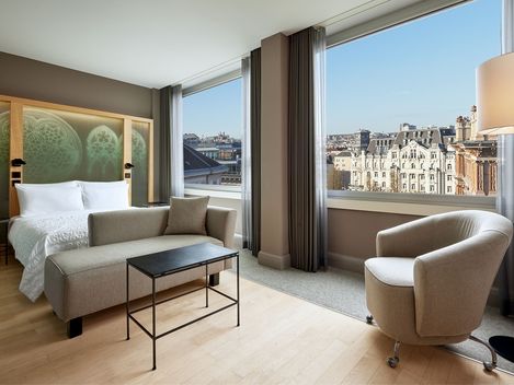 Modernes Hotelzimmer mit Blick auf die Stadt. Mit Bett, Sofa, Sessel und Lampe in der Nähe großer Fenster, die einen Blick auf historische Architektur unter strahlend blauem Himmel bieten.