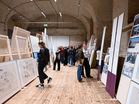 In a large vaulted brick hall with a wooden floor, there are huge paintings on the left and right, which several people are looking at.