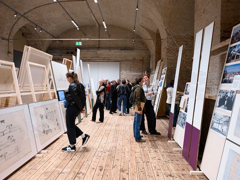 In a large vaulted brick hall with a wooden floor, there are huge paintings on the left and right, which several people are looking at.