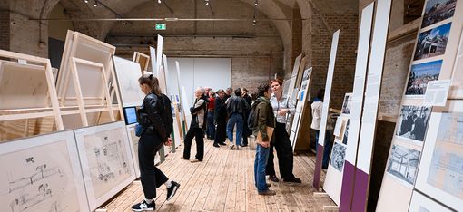 In a large vaulted brick hall with a wooden floor, there are huge paintings on the left and right, which several people are looking at.