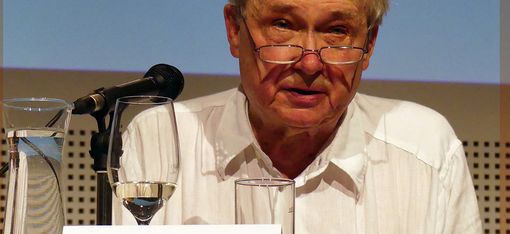 Architekt Hermann Czech, weißes Hemd und Brille sitzt auf der Bühne. Vor ihm steht sein Namensschild und ein Mikrofon.