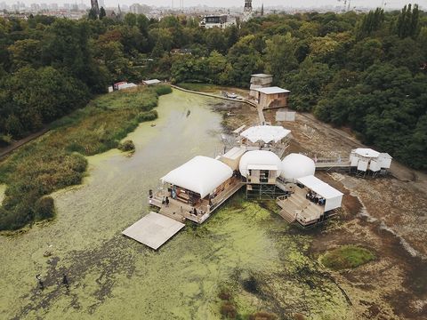 Ein weißes Gebäude, zusammengesetzt aus verschiedenen Raumformen (rund eckig, oval, ...), Terrassen und Stegen, steht teilweise an Land, ein dazugehörender Bauteil schwimmt auf einem Floss in grünem Wasser. Im Hintergrund sieht man den Wald und weit entfernt weitere Häuser.