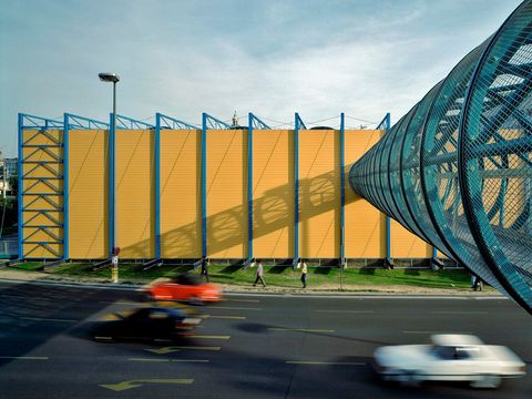 On the right-hand side of the picture, a turquoise tube crosses a busy road to a yellow and turquoise hall-like building on the other side of the road.