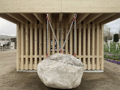 Vor einem Pavillion aus hellem Holz, hängt an einer Kette ein großer Felsen.