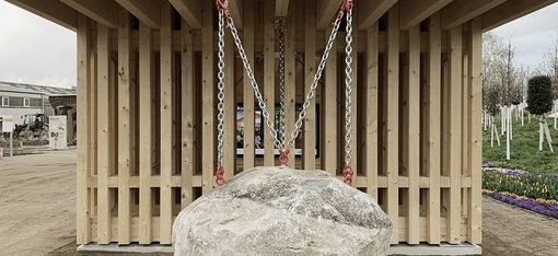 Vor einem Pavillion aus hellem Holz, hängt an einer Kette ein großer Felsen.