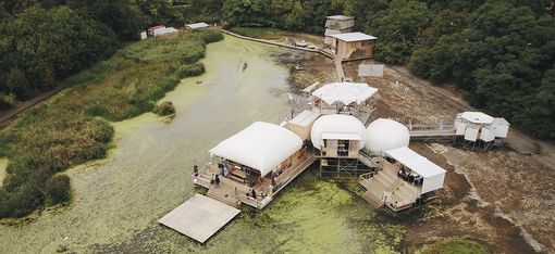 A white building, comprising various shapes (round, square, oval, etc.), terraces and walkways, stands partly on land, whilst an adjoining section floats on a raft in green water. In the background, you can see the forest and, in the distance, other houses.