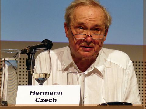 Architect Hermann Czech, wearing a white shirt and glasses, is sitting on stage. In front of him is his nameplate and a microphone.© Foto: Az W