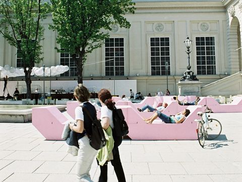 Zwei Personen gehen über den Platz im MuseumsQuartier, im Hintergrund liegen Menschen auf rosa MQ Sitzmöbeln vor einer historischen Gebäudefassade.