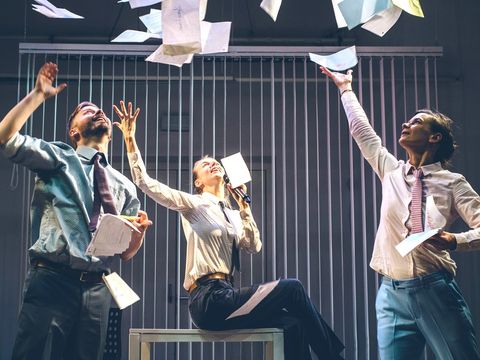 Three people, dressed in smart office attire, are holding lots of pieces of paper, some of which they are throwing into the air. They watch the flying pieces of paper go, smiling.