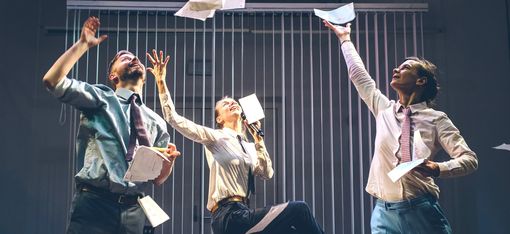 Three people, dressed in smart office attire, are holding lots of pieces of paper, some of which they are throwing into the air. They watch the flying pieces of paper go, smiling.