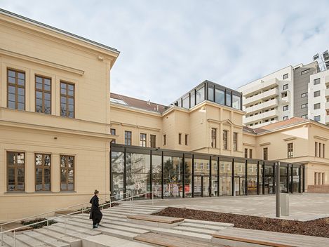 A renovated, single-storey, pale yellow building with a modern steel-and-glass structure in the entrance area and on the roof above the main entrance.