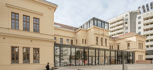 A renovated, single-storey, pale yellow building with a modern steel-and-glass structure in the entrance area and on the roof above the main entrance.